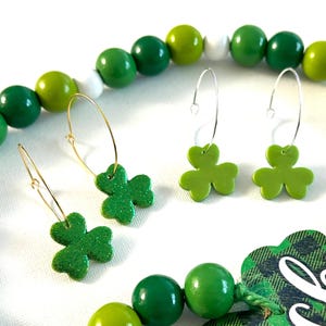 May include: A St. Patrick's Day themed collection featuring green shamrock earrings, a beaded garland, and a decorative tag with the word "Lucky". The earrings have gold and silver hoops. The garland has green, white, and yellow beads. The tag has a green plaid pattern.