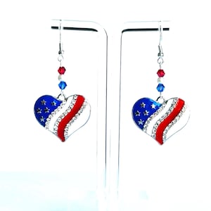 May include: A pair of heart-shaped earrings with a patriotic design. The earrings feature a blue section with white stars, a white section with clear rhinestones, and a red section. The earrings are suspended from silver-colored hooks with red, white, and blue beads.