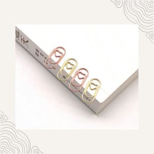 May include: Four metal paper clips in gold and rose gold colours, each with a heart-shaped design. The clips are on the corner of a white book, offering a decorative stationery item.