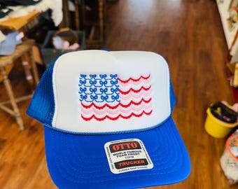 May include: A blue and white trucker hat with a flag design. The hat features a blue brim and mesh back, with a white front panel embroidered with a bow and wave pattern in blue, pink, and red. The hat has a snap closure.