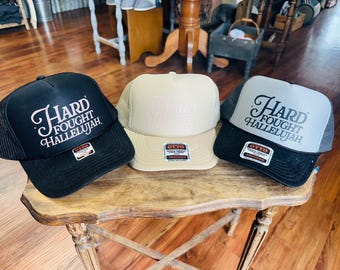 May include: Three trucker hats with the text "Hard Fought Hallelujah" in a script font. The hats are black, tan, and gray, with mesh backs and adjustable snap closures. Each hat has a small label.