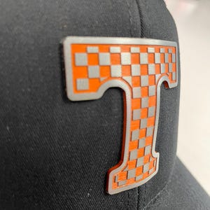 Può includere: Cappellino da baseball nero con un grande emblema a forma di lettera "T" a scacchi arancione e grigio. L'emblema ha un contorno argentato ed è fissato sulla parte anteriore del cappellino. Il cappellino è realizzato in tessuto strutturato.