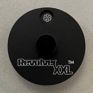 May include: A black, circular object with the text "Revolver XXL" in white. It features a small, circular indentation with a six-circle design and a larger, central cylindrical protrusion.