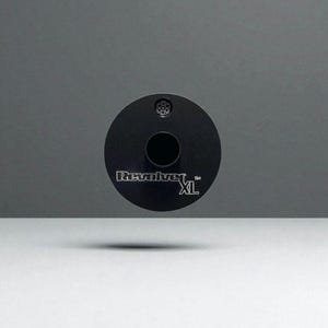 Pode incluir: Um produto preto e circular com o texto "Revolver XL" em branco. O produto tem um orifício central e um padrão circular menor na parte superior. O fundo é uma gradação de cinza e branco.