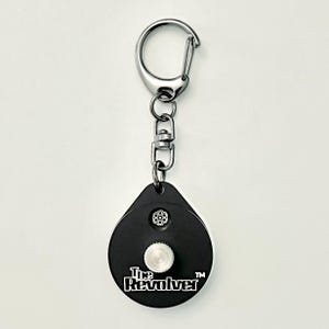 May include: Black and silver keychain with a circular design and the text "The Revolver". The keychain has a silver carabiner clip and a small, round, silver button. The design includes a small, circular pattern.