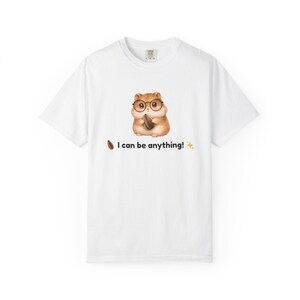 以下が含まれることがあります： メガネをかけたハムスターの漫画が種を持っている白いTシャツ。テキスト「I can be anything！」が下に印刷されています。Tシャツはクルーネックです。