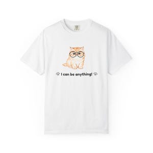 以下が含まれることがあります： 丸いメガネをかけた、薄いオレンジ色の猫のイラストが描かれた白いTシャツ。「I can be anything!」の文字と足跡が猫の下にプリントされています。Tシャツはクルーネックで半袖です。