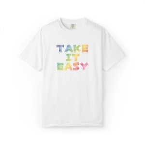 以下が含まれることがあります： 「TAKE IT EASY」の文字がカラフルなピクセルで描かれた白いTシャツ。テキストは青から赤、黄色へと変化する虹色の配色を使用しています。Tシャツはクルーネックです。