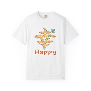 以下が含まれることがあります： 黄色、オレンジ、緑の花柄と青い蝶が描かれた白いTシャツ。「Happy」の文字がオレンジ色のスクリプトでデザインの下に書かれています。Tシャツはクルーネックです。