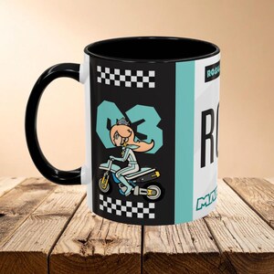 Puede incluir: Taza de cerámica en blanco y negro con un personaje de dibujos animados en una motocicleta. La taza tiene un asa e interior negros, con el nombre del personaje y el número "03" en color turquesa. La taza está sobre una superficie de madera.
