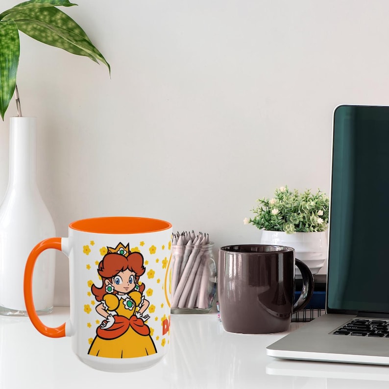 Pu&ograve; includere: Una tazza in ceramica bianca con un bordo e un manico arancioni. La tazza presenta un'immagine di cartone animato della principessa Peach della serie di videogiochi Super Mario Bros. L'immagine mostra la principessa Peach con un vestito giallo con un fiocco rosso e una corona. Lo sfondo della tazza &egrave; bianco con fiori gialli.