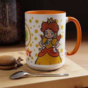 Pu&ograve; includere: Una tazza in ceramica bianca con un bordo e un manico arancioni. La tazza presenta un'immagine di cartone animato della principessa Peach della serie di videogiochi Super Mario Bros. Lo sfondo &egrave; un motivo di fiori gialli e bianchi.