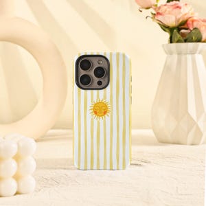 May include: A white and yellow striped phone case featuring a smiling sun graphic. The case has a glossy finish and is designed for a smartphone. The background includes a vase with pink flowers and a decorative object.