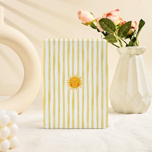 Puede incluir: Un objeto decorativo rectangular con un patrón de rayas amarillas y blancas y una ilustración de un sol sonriente. Se coloca sobre una superficie blanca, con un jarrón blanco y flores rosas en el fondo.