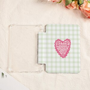 May include: Clear protective case and a light green and white gingham patterned book cover. The book cover has a pink heart with the words "BOOK LOVERS CLUB" inside. The case and cover are on a cream-colored surface.