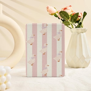 Può includere: Un oggetto rettangolare con un motivo di anatre e strisce verticali rosa e bianche. Le anatre hanno fiocchi blu. L'oggetto è probabilmente una custodia o una cover, forse per un tablet o un e-reader.