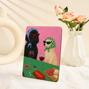 Pu&ograve; includere: Una stampa artistica rettangolare con due cani con occhiali da sole e foulard, che si godono vino e snack. L'opera d'arte ha uno sfondo rosa e un tavolo verde con la parola "SASSY" su una scatola rossa.