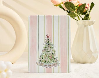 May include: A rectangular Christmas-themed gift box with a watercolor illustration of a decorated Christmas tree. The box has a striped background in pink, white, and green. A red star tops the tree. The box is on a white surface.