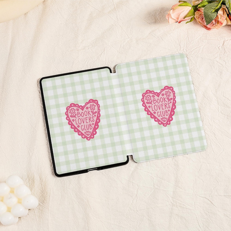May include: An open, black tablet case with a light green and white gingham pattern. Two pink heart-shaped designs with the text "BOOK LOVERS CLUB" are visible on the case. The case is on a cream-colored surface.