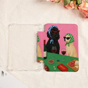 Pu&ograve; includere: Custodia trasparente per telefono con una colorata illustrazione di due cani con occhiali da sole e foulard, che si godono vino e snack. L'opera presenta uno sfondo rosa e un tavolo verde con cibo e la parola "Sassy".