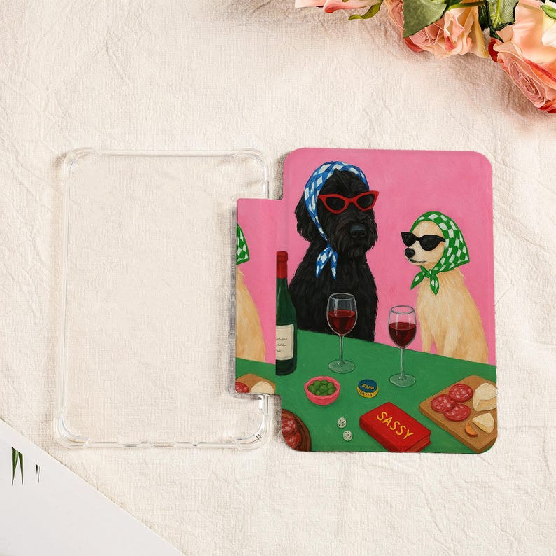 Pu&ograve; includere: Custodia trasparente per telefono con una colorata illustrazione di due cani con occhiali da sole e foulard, che si godono vino e snack. L'opera presenta uno sfondo rosa e un tavolo verde con cibo e la parola "Sassy".