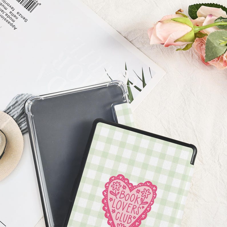 May include: A clear protective case and a tablet case with a green and white gingham pattern. The tablet case features a pink heart with the words "BOOK LOVERS CLUB" and floral accents. The items are on a white surface.