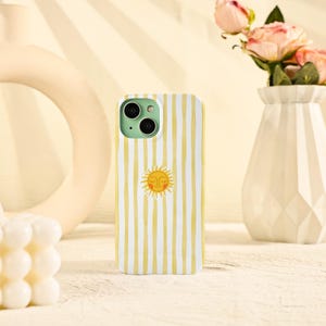 May include: A white and yellow striped phone case featuring a smiling sun graphic. The case has a mint green camera module and is displayed on a light-colored surface. The case is designed to fit a smartphone.