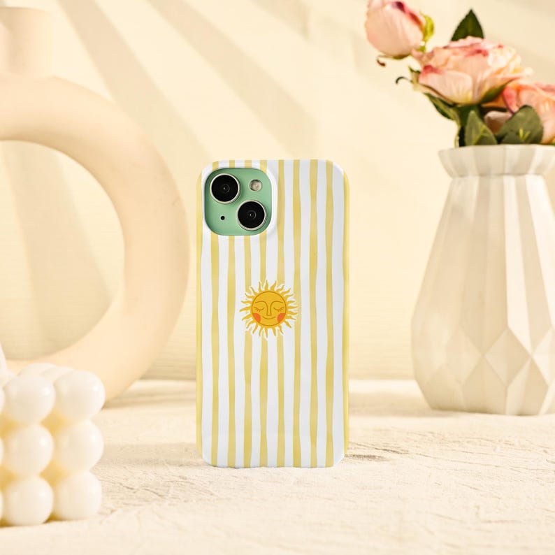 May include: A white and yellow striped phone case featuring a smiling sun graphic. The case has a mint green camera module and is displayed on a light-colored surface. The case is designed to fit a smartphone.
