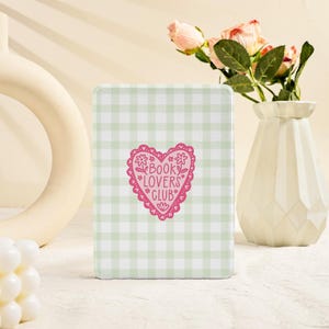May include: A rectangular, light green and white gingham pattern book cover with a pink heart-shaped graphic that reads "BOOK LOVERS CLUB". A white vase with pink roses is in the background.