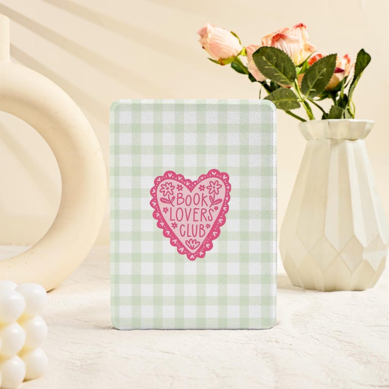 May include: A rectangular, light green and white gingham pattern book cover with a pink heart-shaped graphic that reads "BOOK LOVERS CLUB". A white vase with pink roses is in the background.
