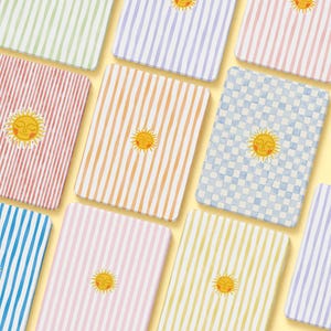 Op de afbeelding: Een verzameling kleurrijke notebooks met patronen. Elk notebook heeft een ander ontwerp, waaronder strepen en ruitpatronen, in tinten groen, paars, roze, oranje, blauw en geel. Op elk notebook staat een lachende zon afgebeeld.