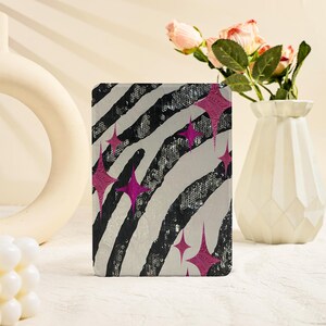 May include: A rectangular tablet case featuring a black and white zebra stripe pattern with a sequined texture. The design is accented with several four-pointed star shapes in a vibrant magenta pink color, scattered across the surface.