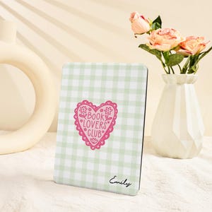 May include: A light green and white gingham pattern e-reader cover with a pink heart-shaped design that reads "Book Lovers Club." The cover also has the name "Emily" printed in cursive. A vase of pink roses is in the background.