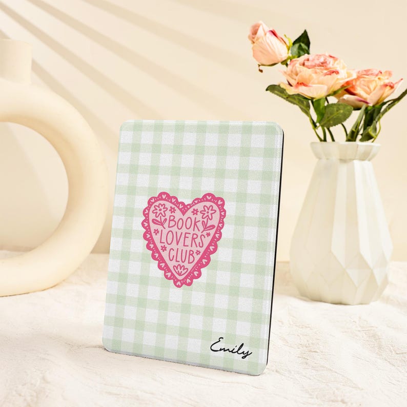 May include: A light green and white gingham pattern e-reader cover with a pink heart-shaped design that reads "Book Lovers Club." The cover also has the name "Emily" printed in cursive. A vase of pink roses is in the background.