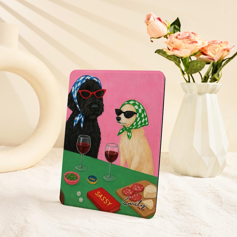 Pu&ograve; includere: Una custodia rosa per tablet con un'illustrazione di due cani con occhiali da sole e foulard, che si godono vino e snack. La scatola rossa dice "Sassy", e il nome "Emily" &egrave; su una tavola di legno.