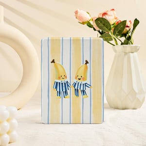 Puede incluir: Un objeto decorativo rectangular con una ilustración caprichosa de dos plátanos antropomórficos con pijamas a rayas azules y blancas. El fondo tiene rayas verticales amarillas y blancas. El objeto está sobre una superficie blanca.