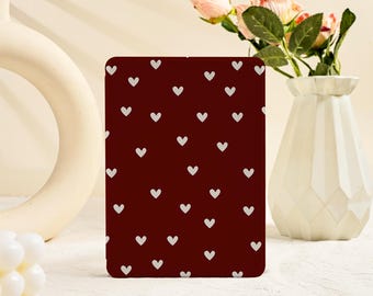 May include: A rectangular item in burgundy, patterned with small, white hearts. It's placed on a white surface, with a white vase holding pink flowers in the background. A white, circular decorative object is also visible.