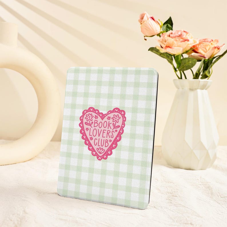 May include: A rectangular, light green and white checkered book cover with a pink heart-shaped graphic that reads "BOOK LOVERS CLUB". The cover is set against a neutral background with a vase of pink roses.