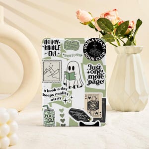 May include: A white Kindle case decorated with various stickers in green and white. The stickers include phrases such as "In My Kindle Era," "Just One More Page," and a ghost reading a book. There is also a "Romantasy Book Club" sticker on the case.