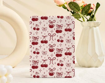 Puede incluir: Una funda rectangular de color crema con un patrón repetido de cerezas rojas con lazos y estrellas. La funda se muestra en posición vertical, mostrando el diseño. Un jarrón blanco con flores rosas está en el fondo.
