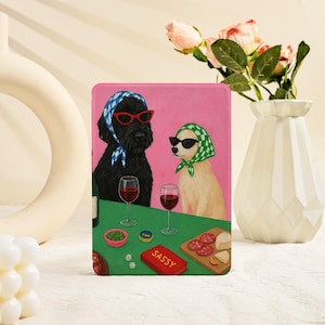 May include: A rectangular tablet case featuring a pink background with a black dog and a tan dog wearing headscarves and sunglasses. They sit at a green table with two wine glasses, a bowl of green olives, a wooden board with salami and cheese, and a red book labeled SASSY.