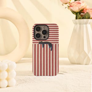 May include: A phone case with a red and white striped pattern and a black cat illustration. The case has a camera cutout and a protective design. The cat is lying down, adding a playful touch to the accessory.