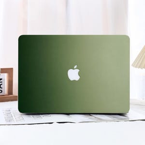 Puede incluir: Un ordenador portátil con una cubierta exterior de color verde degradado liso y sólido. El color transita desde un verde bosque profundo y oscuro en el lado izquierdo hasta un verde oliva más claro y apagado en el derecho, con un logo Apple blanco.