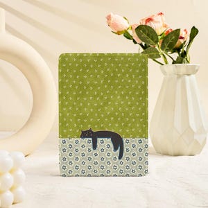 Puede incluir: Objeto rectangular decorativo con una ilustración de gato negro. La mitad superior tiene un fondo verde con detalles florales blancos, mientras que la mitad inferior tiene un patrón floral azul y blanco. Un gato negro está acostado.