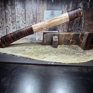 Könnte beinhalten: Eine handgefertigte Axt mit einem Holzgriff, der mit braunem Leder umwickelt ist. Der Axtkopf besteht aus Metall und wird auf einem Holzständer ausgestellt. Der Hintergrund zeigt eine Holzwand und einen Kuhfellteppich.