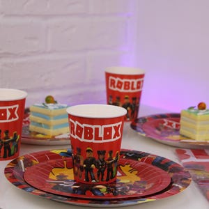 Op de afbeelding: Rode papieren bekers en borden met een Roblox-thema. De bekers en borden zijn voorzien van het Roblox-logo en karakterillustraties. De borden zijn gelaagd met een rood en geel ontwerp. Er zijn ook stukjes cake zichtbaar.