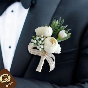 May include: White flower boutonniere with green leaves and a gold ribbon. The boutonniere is pinned to a black suit jacket.