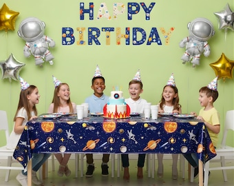 Puede incluir: Una escena de fiesta de cumpleaños con niños, globos y una tarta. La mesa está cubierta con un mantel azul con planetas y cohetes. Las palabras "HAPPY BIRTHAY" están expuestas en la pared. Globos con forma de astronauta y estrella decoran la habitación.