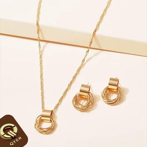 Peut inclure: Un ensemble de bijoux de couleur or comprenant un collier et des boucles d'oreilles assorties. Le collier a une chaîne délicate et un pendentif avec des anneaux entrelacés. Les boucles d'oreilles reprennent le design du pendentif. L'ensemble est présenté sur un fond clair.