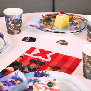May include: A party table setting with Roblox-themed decorations. Includes paper cups, plates, napkins, and a cake roll. The color scheme is red, white, and blue, with cartoon character graphics. A festive arrangement for a birthday celebration.